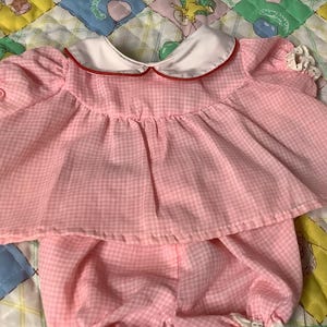 以下が含まれることがあります： 白とピンクのギンガムチェックのベビー人形用ドレス。白い襟と赤いトリムが付いています。ドレスはフリルスカートで、ギンガムチェックのブルマと合わせています。人形はカラフルなパッチワーク模様のキルトの毛布の上に横たわっています。
