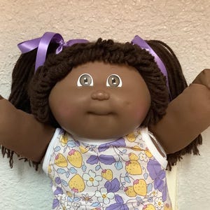 Può includere: Una bambola vintage con pelle scura, capelli castani in codini legati con nastri viola e occhi marroni. La bambola indossa un vestito senza maniche bianco con un motivo floreale di fragole gialle e foglie viola, calzini di pizzo bianchi e scarpe bianche.