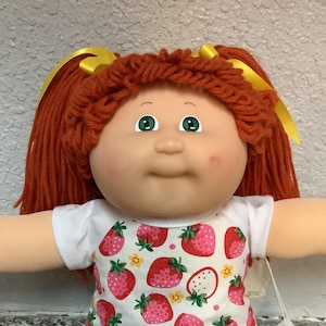 May include: A vintage doll with red yarn hair styled in pigtails with yellow ribbons. The doll has green eyes, rosy cheeks, and is wearing a white outfit with a strawberry print, pink shoes, and white socks.