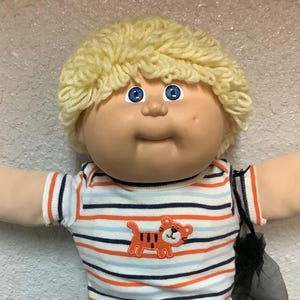 Puede incluir: Muñeca Cabbage Patch Kid con pelo rubio de hilo y ojos azules. La muñeca lleva una camiseta a rayas con un motivo de tigre naranja, pantalones negros y zapatillas blancas con detalles azules. Se ve una bolsa de malla negra.