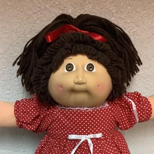 Könnte beinhalten: Vintage Puppe mit braunen Garnhaaren, einem roten Band und einem roten Kleid mit weißen Punkten und weißer Spitzentrimmung. Die Puppe hat ein rundes Gesicht, rosige Wangen und trägt weiße Schuhe mit Spitzentrimmung.