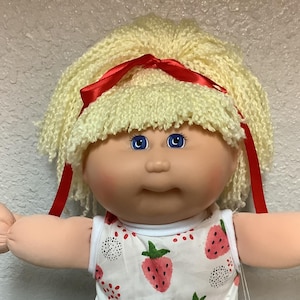 May include: A vintage doll with a blonde, curly wig tied with a red ribbon. The doll has blue eyes and is wearing a white dress and shorts with a strawberry print, lace trim, and red shoes. The doll's arms are outstretched.