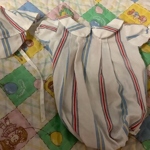 May include: A vintage baby outfit set, comprising a white romper and matching cap. The romper has red and blue vertical stripes. The set is placed on a colourful quilt with cartoon characters and geometric patterns.