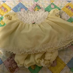 Op de afbeelding: Een gele babyjurk en bijpassende bloomer set. De jurk heeft een geplooide kanten kraag en mouwen, met een klein roze bloemaccent. De bloomers hebben een bijpassende kanten rand. De set wordt gepresenteerd op een kleurrijke patchwork quilt.