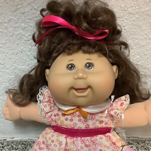May include: A doll with brown curly hair tied with a pink ribbon, featuring a round face and brown eyes. The doll wears a white-collared dress with a pink and orange floral pattern, a pink waist ribbon, orange bows, and a white satin skirt over pink leggings.