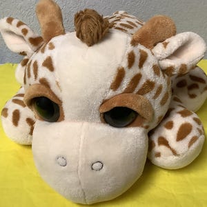 Puede incluir: Un peluche de jirafa con cuerpo blanco y manchas marrones. La jirafa tiene ojos grandes y negros, orejas marrones y pequeños cuernos marrones. El juguete está sobre una superficie amarilla.
