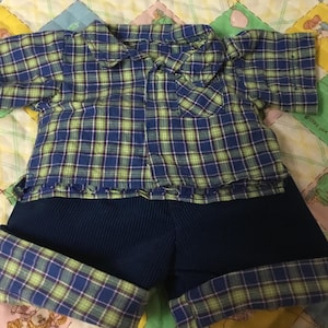 May include: A two-piece baby outfit featuring a blue and green plaid collared shirt and navy blue shorts. The shorts have a ribbed texture and plaid cuffs. The outfit is displayed on a colorful quilt.