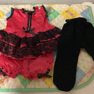 May include: A red satin outfit with black lace trim and bows, consisting of a top and bloomers, alongside black leggings. The outfit is displayed on a colorful, patterned surface.