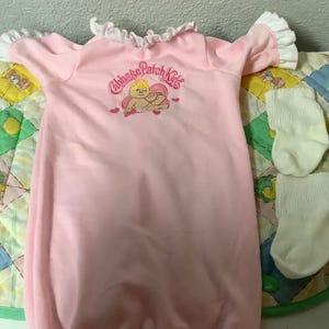 May include: A pink baby gown with white ruffled sleeves and neckline. The gown features a Cabbage Patch Kids graphic and is paired with two white socks. The items are placed on a colorful quilted blanket.