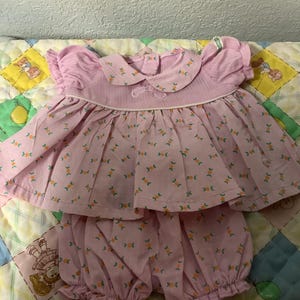 May include: A vintage baby outfit in pink, featuring a top with a Peter Pan collar and matching bloomers. The set is adorned with a repeating floral pattern in orange and green. The outfit is made of a lightweight fabric.