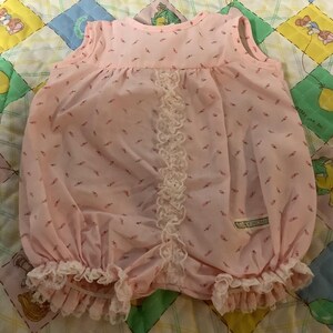 May include: A light pink baby romper with a floral pattern. The romper features a ruffled hem and lace detailing down the front. The garment is sleeveless and has a round neckline. The fabric appears to be lightweight and airy.