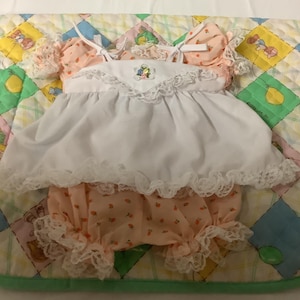 Op de afbeelding: Een witte en roze babypopjurk met kantversiering en een aardbeienprint. De jurk heeft een witte schort-overlay en een bijpassend broekje.