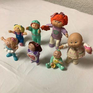 May include: A collection of seven Cabbage Patch Kids figurines. The figurines are all different colours and have different outfits. Some of the figurines are holding accessories, such as a teddy bear and a heart.