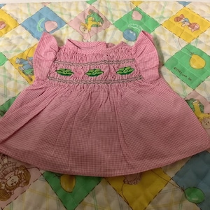 Puede incluir: Vestido de bebé de vichy rosa y blanco con guisantes bordados verdes. El vestido tiene un corpiño fruncido y mangas con volantes.