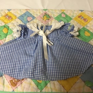 Puede incluir: Un vestido de cuadros azules y blancos con un cuello de encaje blanco y una cinta blanca atada al cuello. El vestido está sobre una tela acolchada de colores.