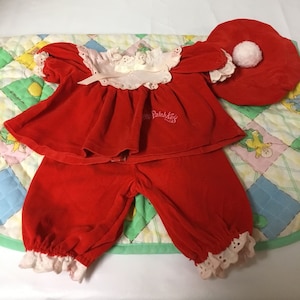 Puede incluir: Conjunto de muñeca de terciopelo rojo con ribete de encaje rosa y un sombrero de terciopelo rojo a juego con un pompón blanco. El conjunto tiene el texto "Little Patch Kids" bordado en la parte delantera.