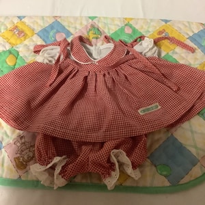 Op de afbeelding: Rode en witte gingham jurk met witte kant en een witte kraag. De jurk heeft een label met de tekst "Pleasant Company".