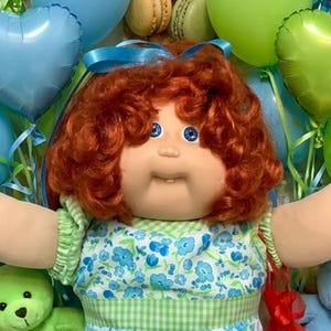 Peut inclure: Une poupée Cabbage Patch Kids aux cheveux roux bouclés portant une robe à fleurs bleue et verte, des chaussettes à bordure en dentelle blanche et des chaussures blanches, entourée d'autres poupées, ours en peluche, ballons en cœur et friandises.