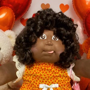 May include: A dark-skinned doll with curly black hair wearing a yellow floral dress with a white ribbon, surrounded by several plush teddy bears, two smaller dolls in heart-patterned outfits, plates of brownies and cookies, and heart-shaped candies.