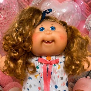 Peut inclure: Une poupée Cabbage Patch Kids aux cheveux bruns bouclés et aux yeux bleus portant une robe blanche à motifs avec un ruban rose, entourée de quatre poupons, deux ours en peluche, des ballons en forme de cœur, des brownies et des bonbons.