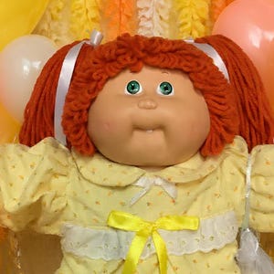May include: A doll with red yarn hair in pigtails, wearing a yellow dress with a white lace collar and yellow ribbon, surrounded by plush teddy bears, heart-shaped balloons, plates of cookies, brownies, and bowls of colorful candies.