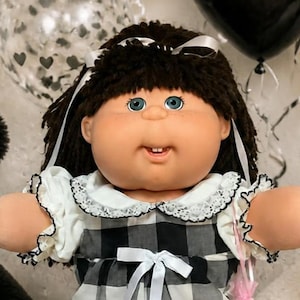 May include: A doll with dark brown hair wearing a black and white plaid dress with a white collar and white lace-trimmed socks and shoes. The doll stands between a black teddy bear and a white teddy bear, with two smaller dolls nearby.
