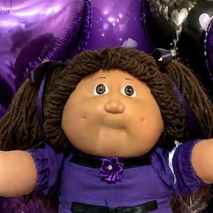 Op de afbeelding: Een grote pop met bruin garen haar in staartjes, gekleed in een paarse jurk met een zwart lint, omringd door kleinere poppen in patronen, paarse hartvormige ballonnen, een bord met chocoladekoekjes en paarse lolly's.