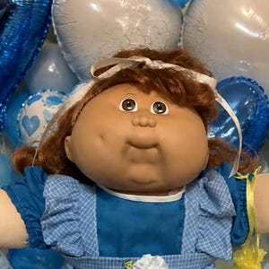 Peut inclure: Une poupée aux cheveux brun roux portant une robe bleue à col blanc et des chaussures blanches, entourée de deux poupées bébés en bleu, deux ours en peluche, des ballons cœurs bleus et blancs, et des biscuits en forme de cœur saupoudrés de bleu.