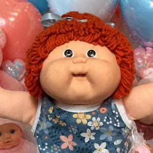 May include: A doll with auburn yarn hair and a blue floral sleeveless dress, wearing light blue ruffled socks. The doll has its arms outstretched and is surrounded by pink and blue heart-shaped balloons, teddy bears, cupcakes, and brownies.