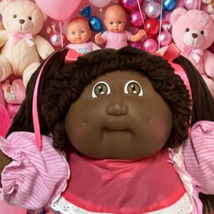 Pode incluir: Uma boneca de pele escura com cabelo de lã castanho escuro em marias-chiquinhas, vestindo um vestido às riscas rosa e branco com um babador rosa, calções com renda branca e sapatos brancos, rodeada por ursos de peluche e balões cor-de-rosa.