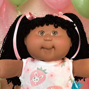 May include: A doll with dark hair in pigtails wearing a white sleeveless dress with a pink strawberry and bunny pattern, white lace-trimmed socks, and white shoes, surrounded by two smaller dolls, two teddy bears, balloons, and candy.