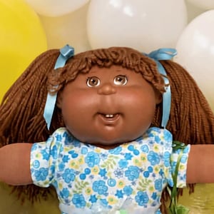 May include: A brown-skinned doll with brown yarn hair in pigtails, wearing a blue and green floral dress with white lace-trimmed boots. The doll is positioned among yellow, white, and blue balloons, two teddy bears, a small doll, and treats.