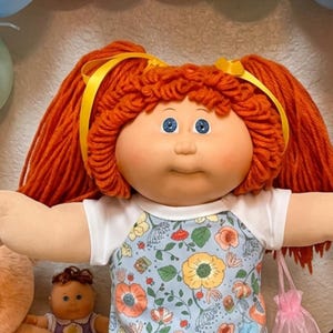 Puede incluir: Una muñeca con cabello de lana naranja brillante en coletas, vistiendo una blusa floral azul y calcetines blancos con volantes, se encuentra entre un gran oso de peluche color melocotón, varias muñecas pequeñas, globos y galletas.