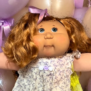 May include: A Cabbage Patch Kids doll with auburn hair and blue eyes wearing a white floral dress with lace trim, white socks with ruffles, and white shoes, flanked by two smaller dolls in lavender outfits amidst purple and cream balloons.