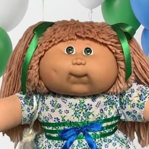May include: A Cabbage Patch Kids doll with brown yarn hair and green eyes, wearing a white dress with a blue floral pattern and a blue ribbon. The doll is surrounded by blue, green, and white balloons, small plush bears, and colorful wrapped candies.