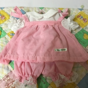 May include: A pink and white gingham dress with a white collar and lace trim. The dress has a matching pair of gingham bloomers with lace trim. The dress is labelled "Our Own Design".