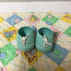 Könnte beinhalten: Ein Paar hellblaue Babyschuhe aus Plastik mit weißen Schnürsenkeln. Die Schuhe liegen auf einer bunten Steppdecke.