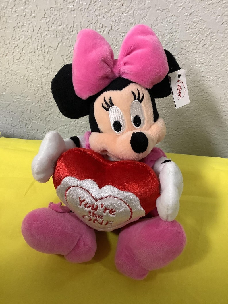 May include: Pink and black plush Minnie Mouse holding a red heart-shaped pillow with the text "You're the One" embroidered on it.