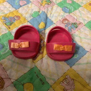 Puede incluir: Un par de zapatos de muñeca de plástico rosa con correas de cinta naranja. Los zapatos están sobre una tela acolchada de colores con un patrón de bebés de dibujos animados y globos.