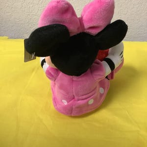 May include: Pink plush Minnie Mouse toy with a black bow and white polka dots on her dress. She is holding a red heart in her hands.