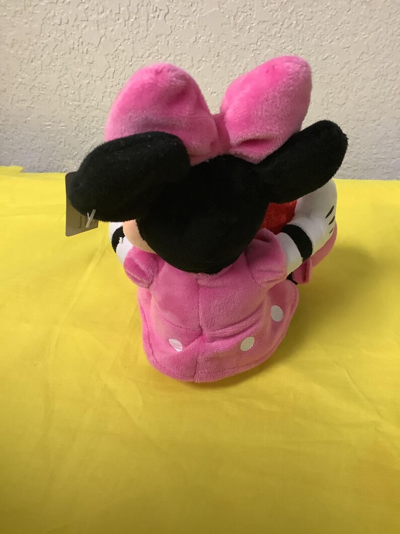 May include: Pink plush Minnie Mouse toy with a black bow and white polka dots on her dress. She is holding a red heart in her hands.