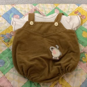 Puede incluir: Un pelele de bebé de pana marrón con una camiseta blanca debajo. El pelele tiene tirantes con botones y un pequeño aplique de elefante. El conjunto se muestra sobre un edredón de retazos colorido.