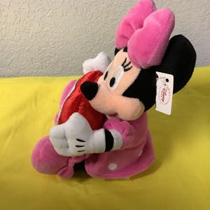 May include: Pink plush Minnie Mouse holding a red heart. She is wearing a pink dress with white polka dots and a pink bow. The plush toy has a Disney Store tag.