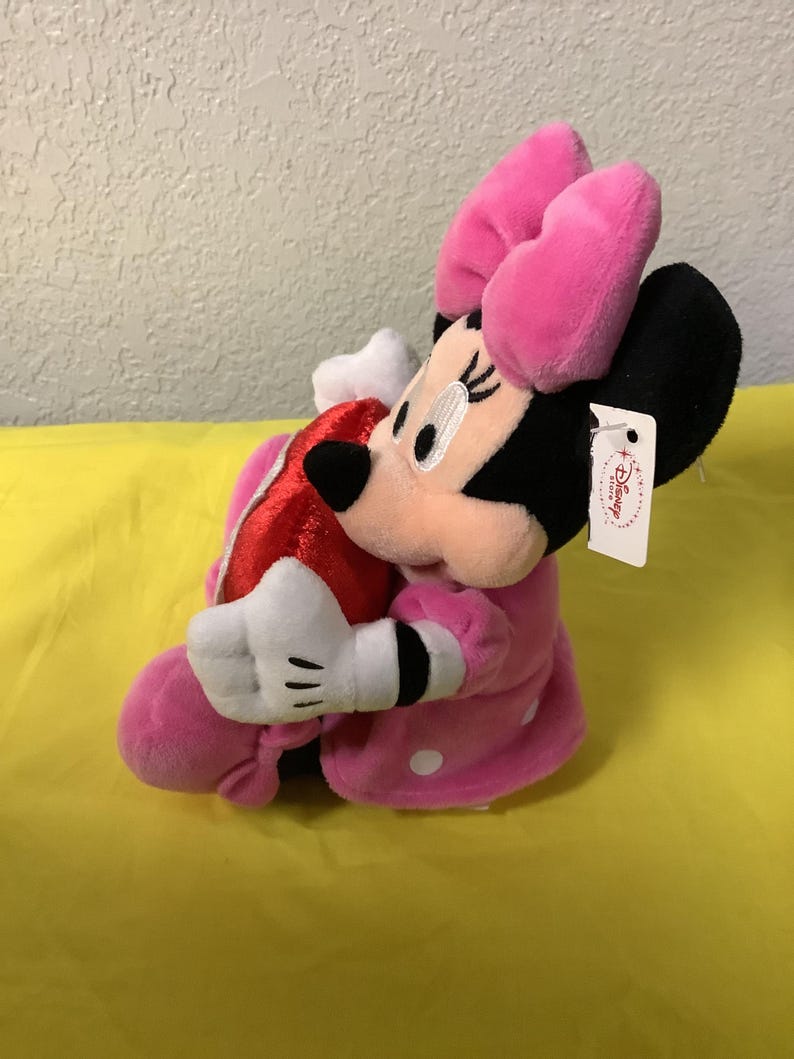 May include: Pink plush Minnie Mouse holding a red heart. She is wearing a pink dress with white polka dots and a pink bow. The plush toy has a Disney Store tag.