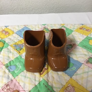 May include: A pair of brown plastic cowboy boots for a doll. The boots are on a quilted fabric with a colourful pattern.