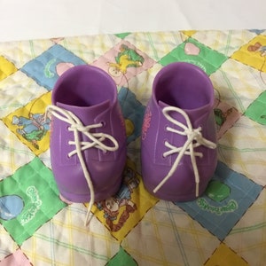 May include: A pair of purple doll shoes with white laces. The shoes have a floral design and are made of plastic.