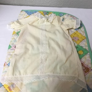 May include: A pale yellow baby gown with short sleeves and a white lace collar and trim. The gown is laid on a colorful patchwork quilt with a green border. The quilt features various cartoon characters.