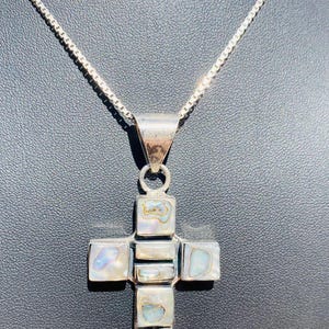 Può includere: Un ciondolo a croce in argento con inserti in conchiglia di abalone iridescente. La croce è composta da diversi segmenti rettangolari e quadrati di conchiglia incastonati in una cornice in argento lucido, appesa a una sottile catenina d'argento.