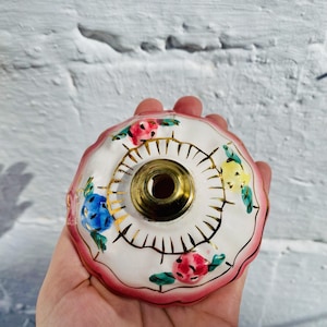Pode incluir: Um acabamento de lâmpada de cerâmica branca com uma borda rosa e detalhes dourados. O acabamento é decorado com flores pintadas em tons de rosa, azul, amarelo e verde.