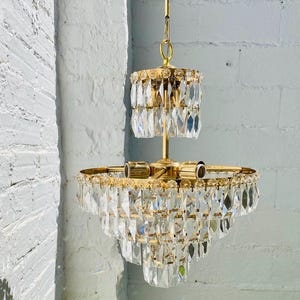 May include: A two-tier chandelier featuring a polished gold-tone metal frame and numerous clear, faceted glass crystals. The fixture includes a central rod, a circular top tier, and a wider circular bottom tier, all adorned with hanging crystal prisms.